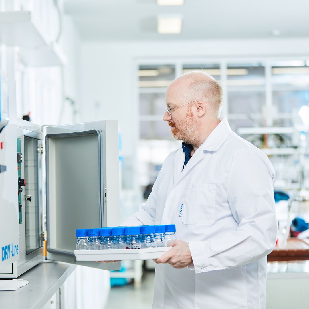 Ein Labortechniker in einem weißen Laborkittel trägt ein Tablett mit Fläschchen mit blauen Kappen zu einem Schrank in einem hellen, sauberen Laborraum.