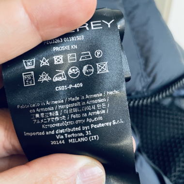 Eine Hand hält ein Pflegeetikett für ein Kleidungsstück mit Wäschesymbolen und mehrsprachigem Text, der angibt, dass der Artikel in Armenien hergestellt wurde.