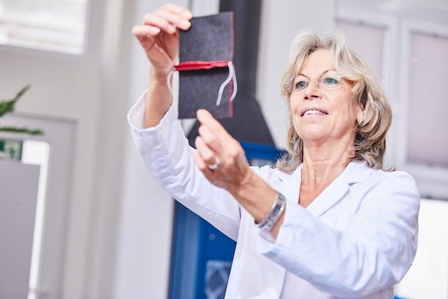 Eine Person in einem weißen Laborkittel hält ein kleines schwarzes, mit roter Schnur verschnürtes Büchlein hoch und demonstriert es in einem hellen Innenraum.