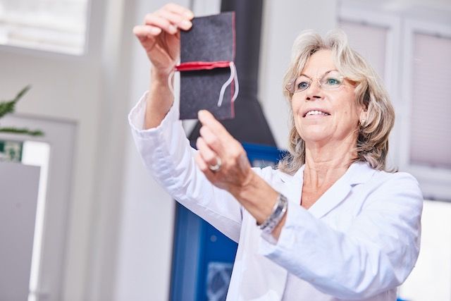 Eine Person in einem weißen Laborkittel hält ein kleines schwarzes, mit roter Schnur verschnürtes Büchlein hoch und demonstriert es in einem hellen Innenraum.