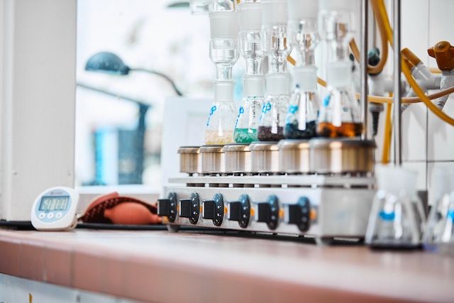 Ein Laboraufbau mit Glaskolben mit gefärbten Flüssigkeiten auf einem beheizten Sockel, einer Zeitschaltuhr und Knöpfen für Einstellungen.