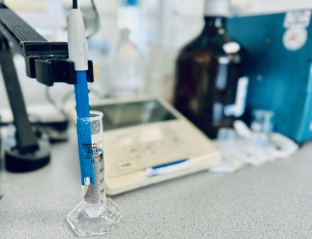 Eine Laboreinrichtung mit einem Messzylinder mit blauer Sonde, einer Digitalwaage und verschiedenen Laborgeräten im Hintergrund.