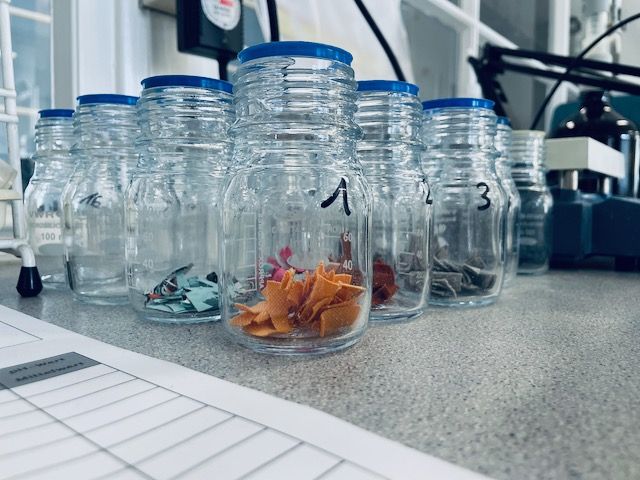 Sieben durchsichtige Gläser mit blauen Deckeln, die mit Nummern beschriftet sind, enthalten bunte Stoffreste, die ordentlich auf einer Arbeitsfläche angeordnet sind.