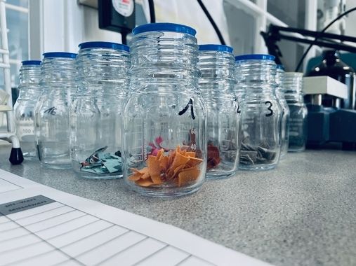 Auf einer grauen Tischplatte steht neben Papierkram eine Reihe von Klarsichtgläsern, gefüllt mit bunten, mit Nummern versehenen Stoffresten.