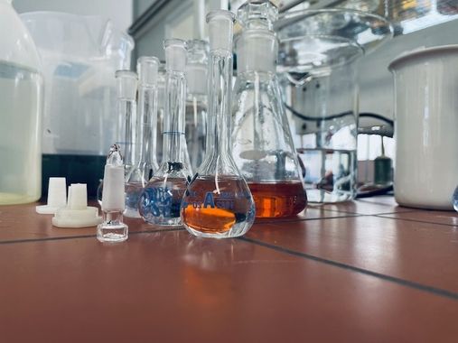 Eine Sammlung verschiedener Laborgeräte aus Glas, darunter Kolben und Bechergläser, gefüllt mit bunten Flüssigkeiten auf einer gefliesten Oberfläche.